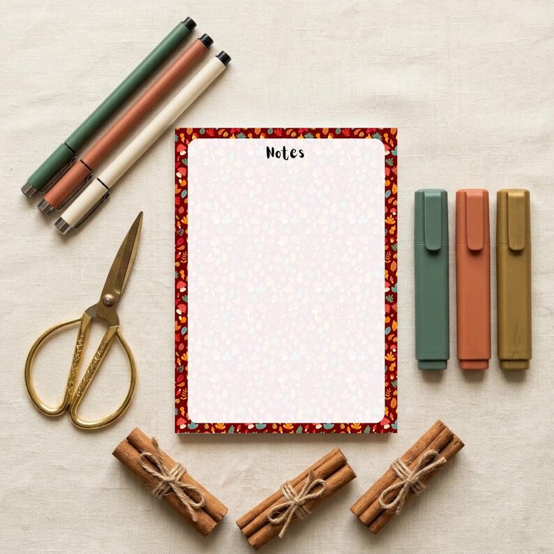 May include: A rectangular notepad with a white center and a border featuring a pattern of small autumn leaves in red, orange, and green. The word Notes is printed at the top. Included are three pens, three markers, gold scissors, and three cinnamon sticks.