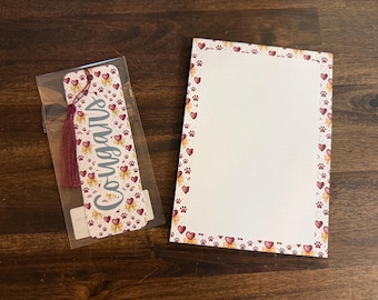 May include: A bookmark in a clear plastic sleeve with a maroon tassel and the word Cougars printed on it, alongside a rectangular notepad. Both items feature a repeating pattern of small maroon hearts, yellow bows, and paw prints.