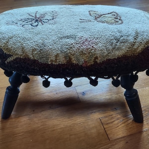 May include: A vintage footstool with a round, tufted top featuring a floral design and two butterflies. The stool has a black wooden base with turned legs and a black tassel fringe around the top.