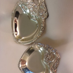 May include: Two silver heart-shaped serving dishes with ornate floral designs and handles. The dishes are made of polished silver and have a reflective surface.