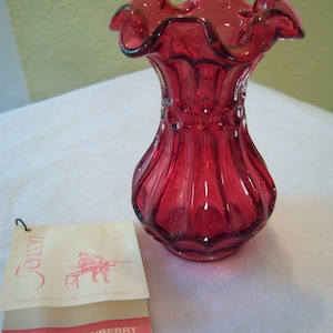 May include: A red glass vase with a ruffled top and a textured body. The vase is handcrafted with pure gold and is labelled "Country Cranberry".