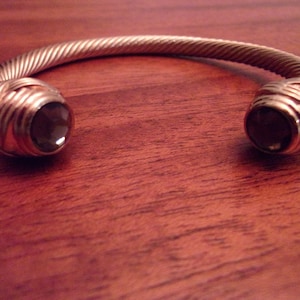 Puede incluir: Un brazalete de plata con un diseño trenzado y dos piedras de color marrón oscuro.