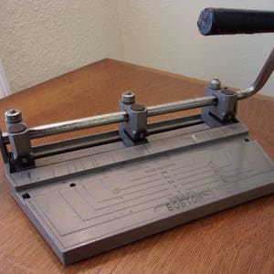 May include: A silver metal hole punch with a black handle. The punch has a metal bar that runs across the top and a series of holes for different sizes of punches. The punch is labeled "Hole Selector" and "Boston".
