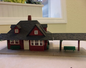 May include: A miniature model of a train station. The building is painted red with a dark gray roof and a brick chimney. It has white window frames and a small covered platform with a green bench.