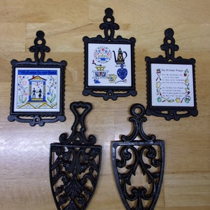 Può includere: Set di cinque sottopentola in ghisa nera con disegni decorativi. Due sottopentola hanno una piastrella quadrata con un design colorato. Una piastrella presenta una casa con il testo "Benvenuti a casa nostra". L'altra piastrella presenta una scena da cucina con un macinacaffè, una teiera e altri oggetti da cucina. Gli altri tre sottopentola hanno la forma di un ovale appuntito con un design floreale decorativo.