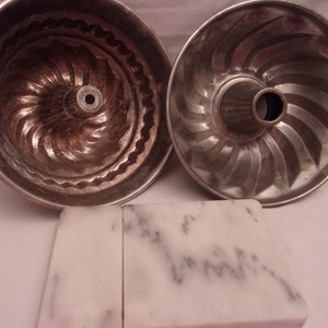 Op de afbeelding: Twee zilveren metalen bundt cakevormen met een swirl-ontwerp. De vormen liggen op twee witte marmeren tegels.