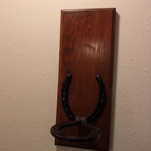 Op de afbeelding: Een houten, aan de muur gemonteerde hoefijzerhouder met een zwart hoefijzer. Het hoefijzer is met schroeven aan het hout bevestigd en heeft een afgeronde onderkant.