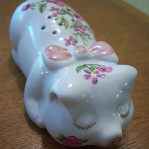 Könnte beinhalten: Ein weißes Keramik-Sparschwein mit rosa Blumenmotiven und einer rosa Schleife. Das Sparschwein hat einen geschlossenen Mund und ist mit rosa und lila Blumen verziert.