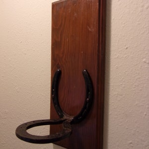 Op de afbeelding: Een houten wandhaak in de vorm van een hoefijzer. Het hoefijzer is van zwart metaal en is gemonteerd op een donkerbruine houten plaat.
