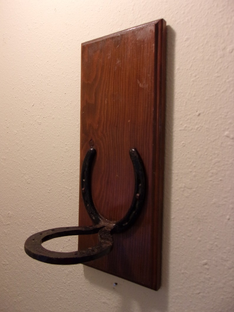 Op de afbeelding: Een houten wandhaak in de vorm van een hoefijzer. Het hoefijzer is van zwart metaal en is gemonteerd op een donkerbruine houten plaat.