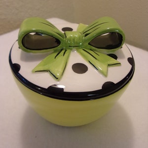 May include: A yellow ceramic container with a white lid decorated with black polka dots and a green bow.