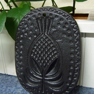 May include: Black cast iron pineapple-shaped trivet. The trivet has an oval shape with a detailed pineapple design in the center and a dotted border. A small hole is at the top for hanging. The trivet is likely used for placing hot cookware on a table.
