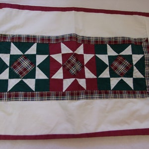 Könnte beinhalten: Ein weißer Tischläufer mit rotem Rand und einem Patchwork-Muster mit grünen, roten und weißen Sternen. Die Sterne sind von einem karierten Rand umgeben.