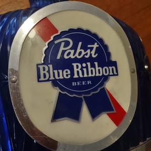 May include: A vintage Pabst Blue Ribbon beer tap handle. The handle is a deep blue color with a circular emblem featuring the Pabst Blue Ribbon logo in blue and white, with a red accent. The handle has a silver-colored metal trim.