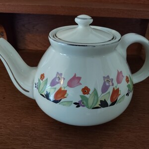 May include: White teapot with a floral design featuring pink, orange, purple, and green flowers. The teapot has a silver trim around the lid and spout.