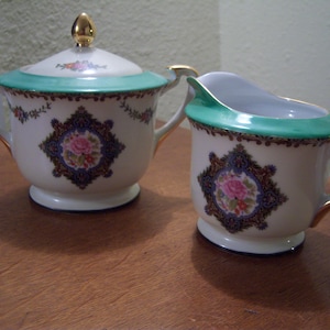 Puede incluir: Un juego de azucarero y jarra de crema de porcelana blanca con ribetes dorados y diseños florales. El azucarero tiene una tapa con un pomo dorado. La jarra de crema tiene un asa.