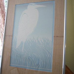 May include: A framed print of a white heron standing in a blue and white field of grass. The frame is made of wood with a light brown finish and has a silver metal border.