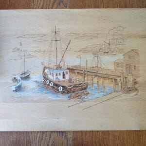 May include: A wooden art piece depicting a coastal scene with boats, a pier, and a lighthouse. The artwork features a detailed illustration of fishing boats, a pier, and a small building, all set against a light-colored wooden background.