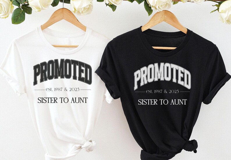 May include: Two t-shirts, one white and one black, each with the word "PROMOTED" in a collegiate-style font. Below the word is "est. 1987 & 2023" and "SISTER TO AUNT". The shirts hang on wooden hangers.