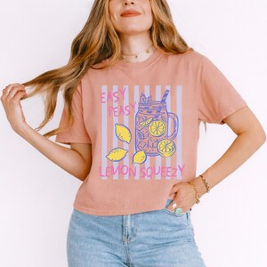 Pu&ograve; includere: T-shirt color pesca con grafica di un barattolo pieno di limoni e ghiaccio, con la scritta "EASY PEASY LEMON SQUEEZY" in viola e giallo. La maglietta ha maniche corte e girocollo.