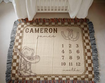 May include: A rectangular woven throw blanket with a beige center and a brown and beige checkered border. The blanket features the name Cameron James, a cowboy hat, a pair of cowboy boots, a horseshoe, and the numbers 1 through 12 for months.