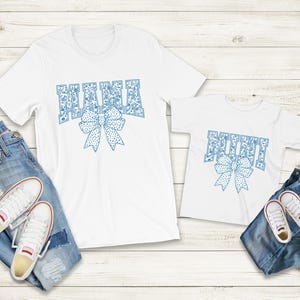 May include: Two white t-shirts with blue patterned text. The larger shirt says "Mama" with a blue bow. The smaller shirt says "Mini" with a blue bow.