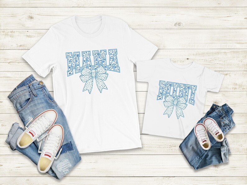 May include: Two white t-shirts with blue patterned text. The larger shirt says "Mama" with a blue bow. The smaller shirt says "Mini" with a blue bow.