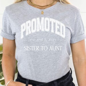 May include: A light gray t-shirt with the word "PROMOTED" in white arched lettering. Below the word "PROMOTED" is the text "est. 1987 & 2025" and below that is the text "SISTER TO AUNT". The shirt has rolled-up sleeves.