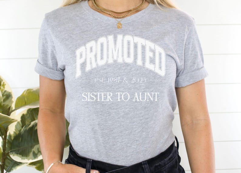 May include: A light gray t-shirt with the word "PROMOTED" in white arched lettering. Below the word "PROMOTED" is the text "est. 1987 & 2025" and below that is the text "SISTER TO AUNT". The shirt has rolled-up sleeves.