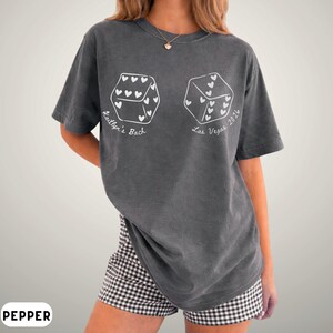 May include: A gray t-shirt with a dice graphic. Each die face has heart shapes. The text "Kaitlyn's Bach" and "Las Vegas 2024" are printed on the shirt. The model is wearing black and white checkered shorts.