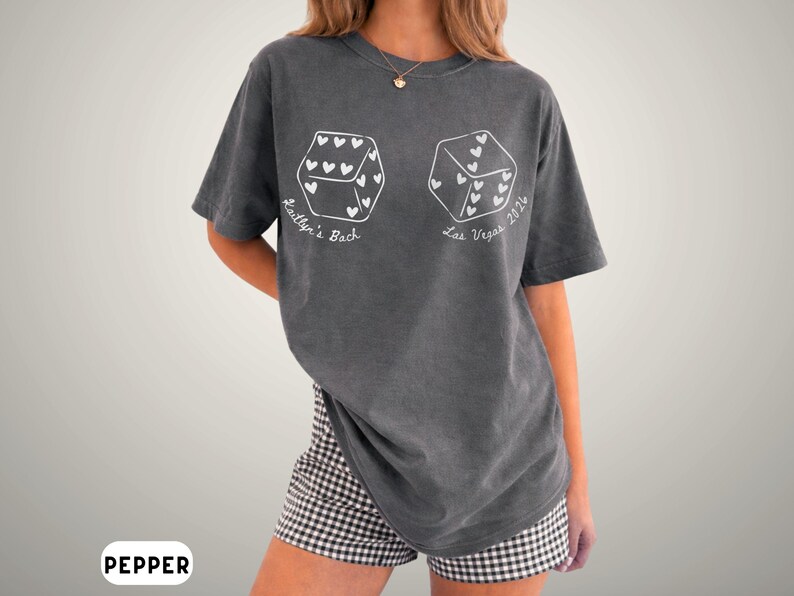 May include: A gray t-shirt with a dice graphic. Each die face has heart shapes. The text "Kaitlyn's Bach" and "Las Vegas 2024" are printed on the shirt. The model is wearing black and white checkered shorts.
