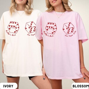 May include: Ivory and blossom-colored t-shirts with a dice graphic. Each die features heart-shaped pips and the text "Kaitlyn's Bach Las Vegas 2024" in red. The shirts are a casual, oversized fit.