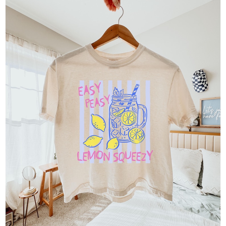 Pu&ograve; includere: T-shirt beige con un disegno di limonata con un barattolo, limoni e la scritta "Easy Peasy Lemon Squeezy" in rosa e blu. La maglietta &egrave; appesa a una gruccia di legno.