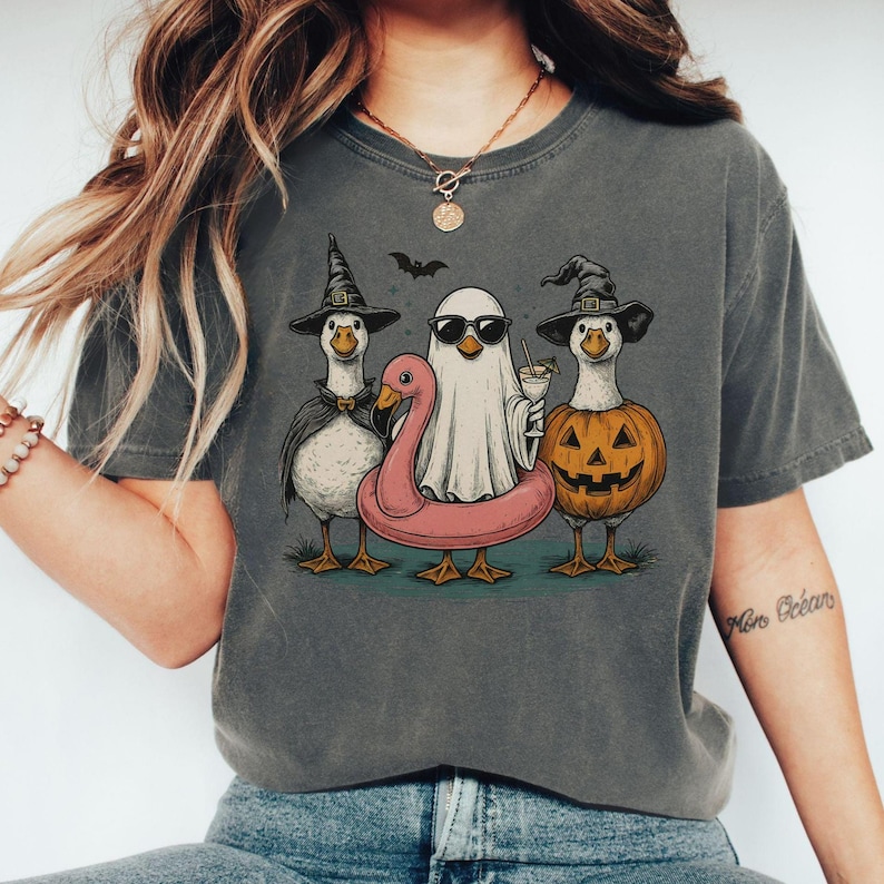 May include: Dark gray t-shirt featuring a Halloween-themed graphic. The design includes three geese dressed as a witch, a ghost, and a jack-o'-lantern, with a pink flamingo floatie. A bat flies above them. The geese are standing on a green surface.