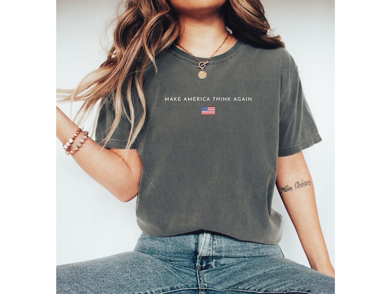 May include: Dark gray t-shirt with the text "MAKE AMERICA THINK AGAIN" and a small American flag graphic. The shirt is a comfortable, casual style, suitable for everyday wear.