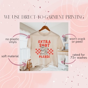 K&ouml;nnte beinhalten: Cremefarbenes T-Shirt mit den roten Worten "EXTRA SHOT PLEASE" und einer Kaffeemaschinen-Grafik. Das Shirt h&auml;ngt an einem Holzb&uuml;gel. Der Text "WE USE DIRECT-TO-GARMENT PRINTING" steht oben.