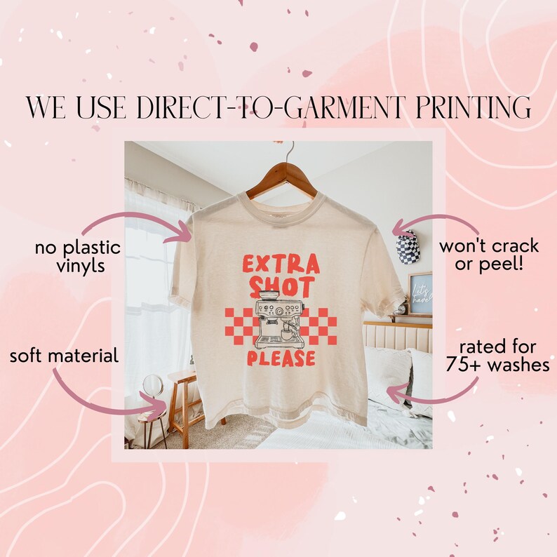 K&ouml;nnte beinhalten: Cremefarbenes T-Shirt mit den roten Worten "EXTRA SHOT PLEASE" und einer Kaffeemaschinen-Grafik. Das Shirt h&auml;ngt an einem Holzb&uuml;gel. Der Text "WE USE DIRECT-TO-GARMENT PRINTING" steht oben.
