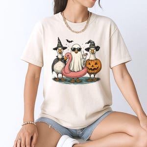 May include: Cream-colored t-shirt featuring a Halloween-themed graphic. The design showcases three geese dressed in costumes: a witch, a ghost, and a jack-o'-lantern. A pink flamingo floatie and a bat are also included in the illustration.