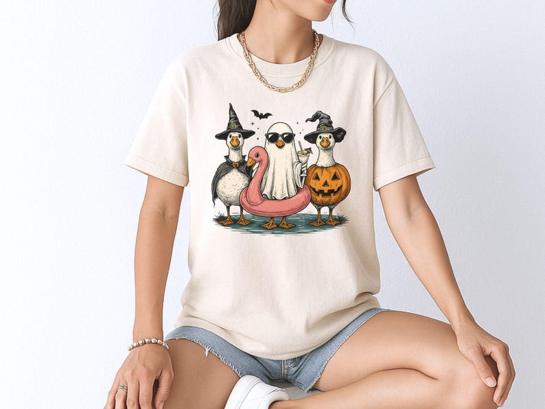 May include: Cream-colored t-shirt featuring a Halloween-themed graphic. The design showcases three geese dressed in costumes: a witch, a ghost, and a jack-o'-lantern. A pink flamingo floatie and a bat are also included in the illustration.