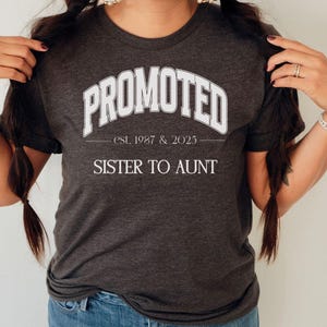 May include: Dark gray t-shirt with the text "PROMOTED" in white, arched across the top. Below, it reads "est. 1987 & 2023" and "SISTER TO AUNT" in white. The shirt is a crew neck style.