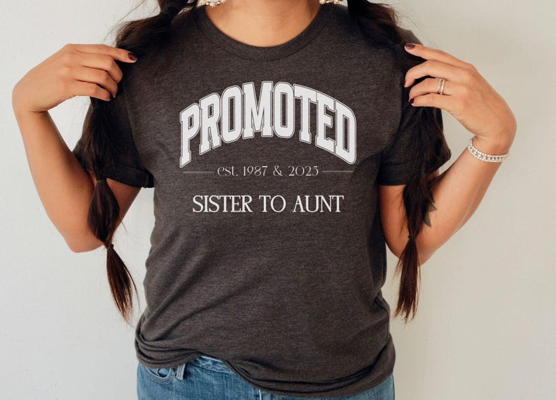 May include: Dark gray t-shirt with the text "PROMOTED" in white, arched across the top. Below, it reads "est. 1987 & 2023" and "SISTER TO AUNT" in white. The shirt is a crew neck style.