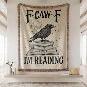 May include: Beige woven throw blanket with a black raven illustration perched on books, with the text "F-CAW-F I'M READING". The blanket has a fringed edge and is displayed on a wooden shelf.