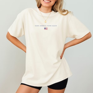 May include: Cream-colored t-shirt with the text "MAKE AMERICA THINK AGAIN" in navy blue, along with a small American flag graphic. The shirt is a relaxed fit.