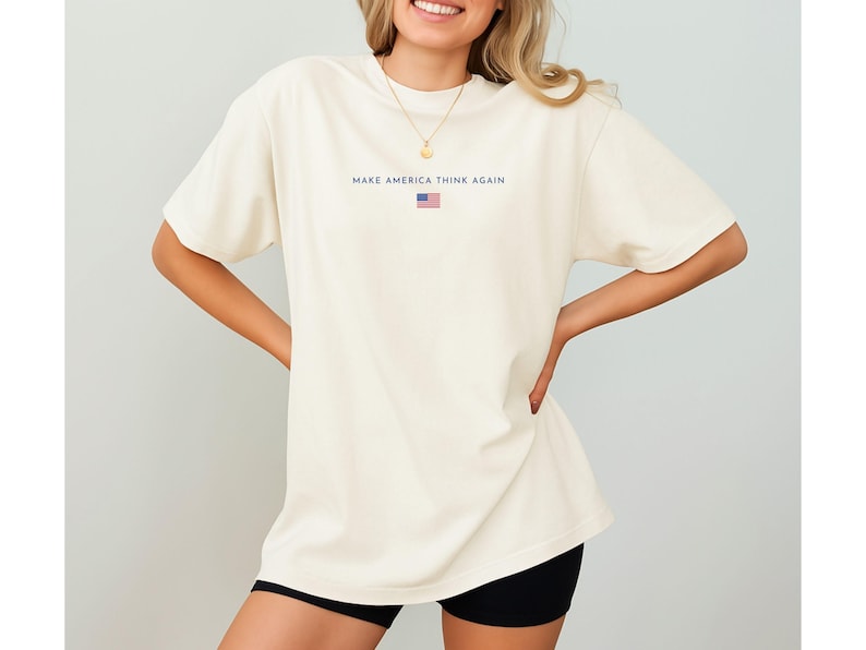 May include: Cream-colored t-shirt with the text "MAKE AMERICA THINK AGAIN" in navy blue, along with a small American flag graphic. The shirt is a relaxed fit.