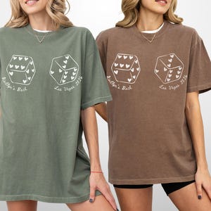 May include: Two oversized t-shirts, one olive green and one brown, each featuring a white graphic of two dice with heart-shaped pips. The text "Kaitlyn's Back" and "Las Vegas 2024" are printed below the dice.
