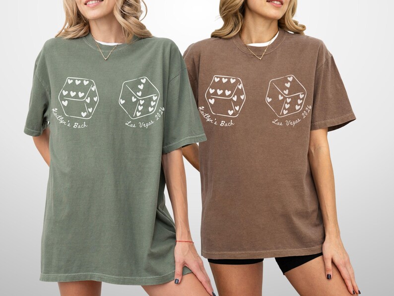 May include: Two oversized t-shirts, one olive green and one brown, each featuring a white graphic of two dice with heart-shaped pips. The text "Kaitlyn's Back" and "Las Vegas 2024" are printed below the dice.