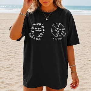 May include: Black t-shirt with white dice graphics, each featuring heart-shaped pips. Text on the shirt reads "Kaitlyn's Bach" and "Las Vegas 2026". The shirt is worn on a beach.