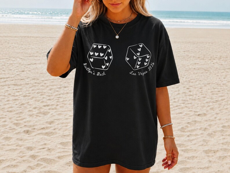May include: Black t-shirt with white dice graphics, each featuring heart-shaped pips. Text on the shirt reads "Kaitlyn's Bach" and "Las Vegas 2026". The shirt is worn on a beach.