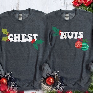 May include: Two gray sweatshirts with a Christmas theme. One sweatshirt says "CHEST" with a holly sprig and the other says "NUTS" with a holly sprig and two Christmas ornaments.