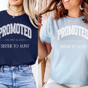 May include: Two t-shirts, one navy blue and one light blue, each with the word "PROMOTED" in a white, arched font. Below, the text reads "est. 1987 & 2025" and "SISTER TO AUNT". The shirts are casual, with rolled sleeves.